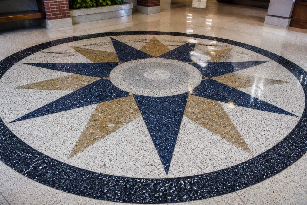 Close-up of a decorative terrazzo floor featuring a custom medallion design with school colors, marble aggregate and glass aggregate visible in the polished surface under bright institutional lighting