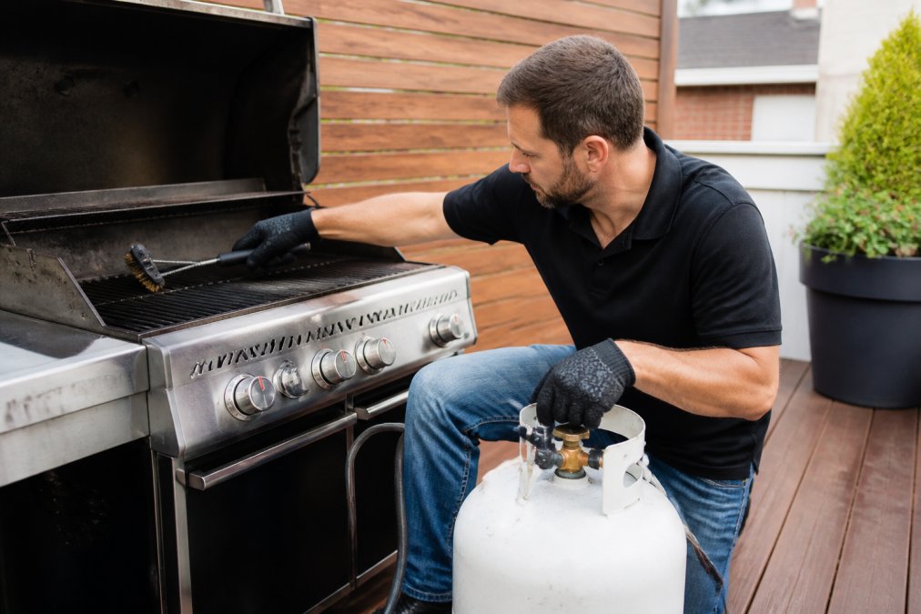 Homeowner cleaning grill grates and checking propane tank connections during routine backyard grill maintenance, wearing work gloves with tools on a wooden deck