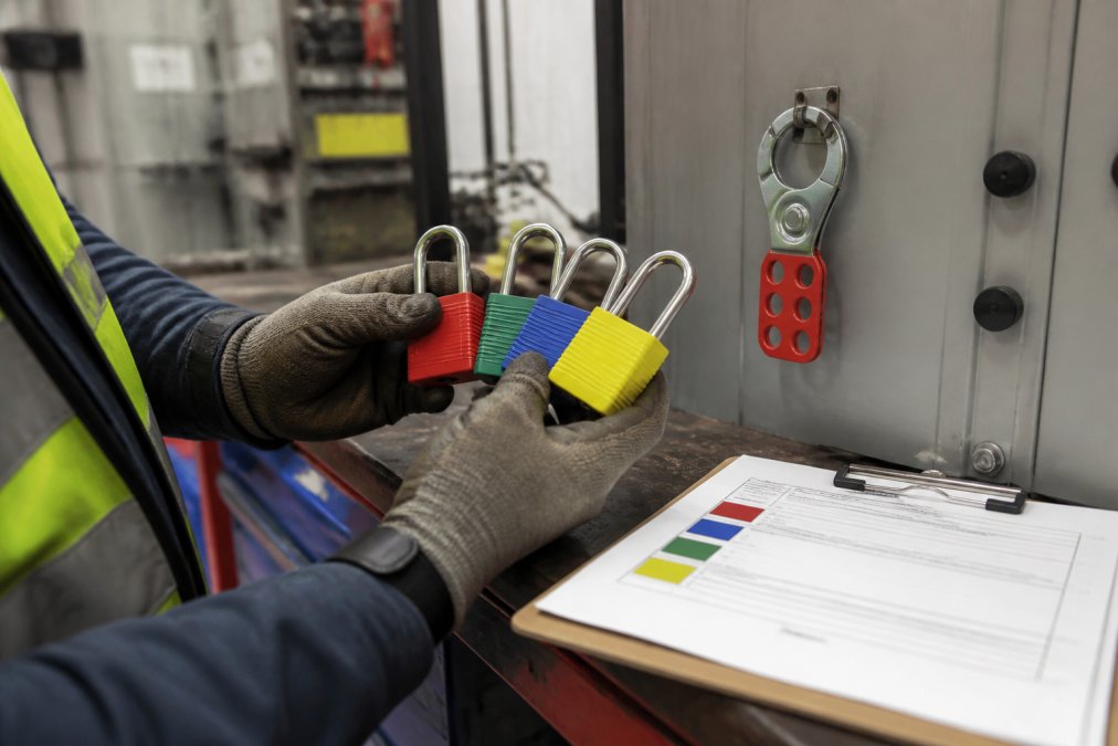 A safety manager's gloved hands holding several lockout padlocks in different colors (red, green, blue, yellow) next to a steel hasp on industrial equipment, with a clipboard of color-coded lockout tags visible on a workbench in a maintenance shop