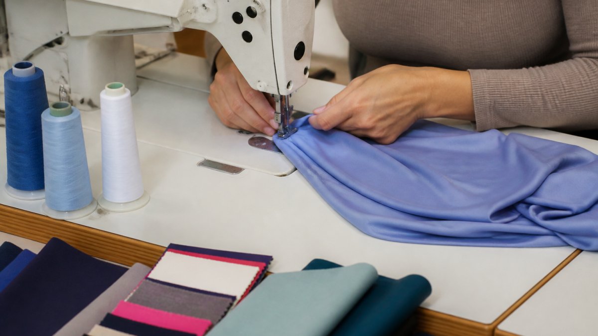 Close-up of a seamstress's hands guiding a stretchy polyester knit fabric under a sewing machine needle, with spools of thread and fabric swatches nearby on a work table