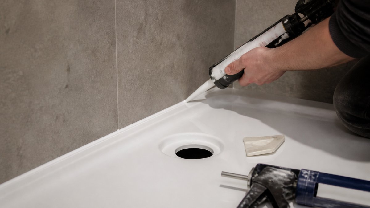 Close-up of a person applying a smooth bead of white silicone sealant along the base of a bathroom wall panel where it meets a shower tray, with a smoothing tool and cartridge gun visible
