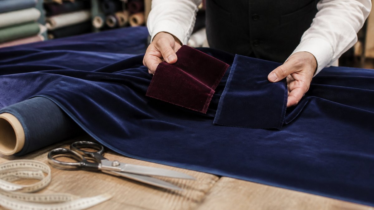 Close-up of a tailor's hands holding two velvet fabric swatches side by side over a worktable, with a measuring tape and shears nearby in a bright studio