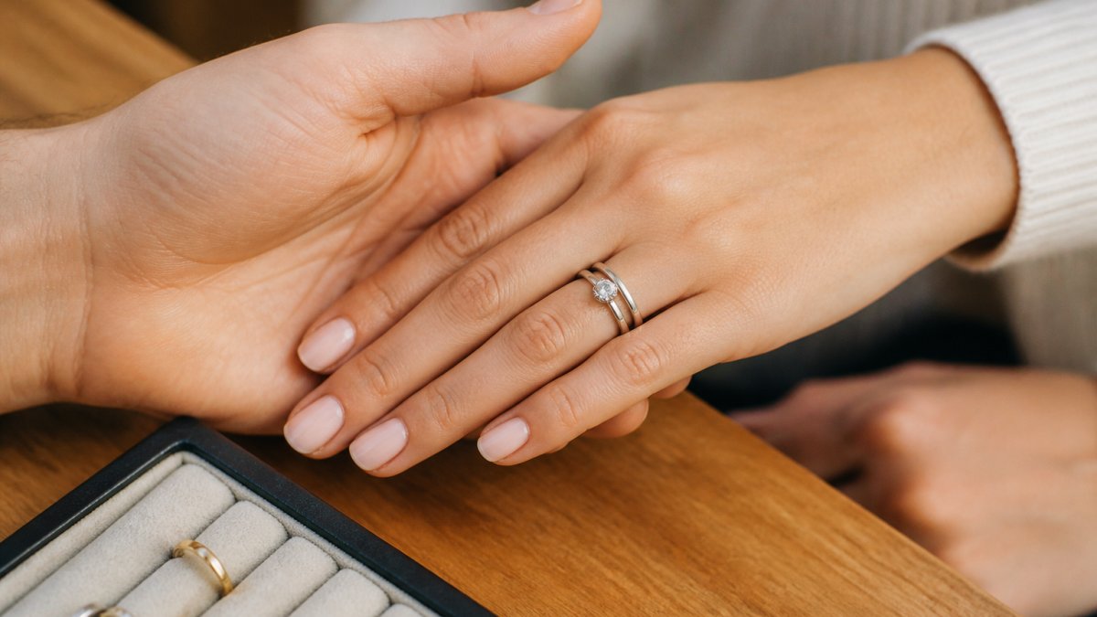 Close-up of a couple's hands together, one wearing an engagement ring and a contoured wedding band sitting flush side by side, soft natural light on a wooden jeweller's counter