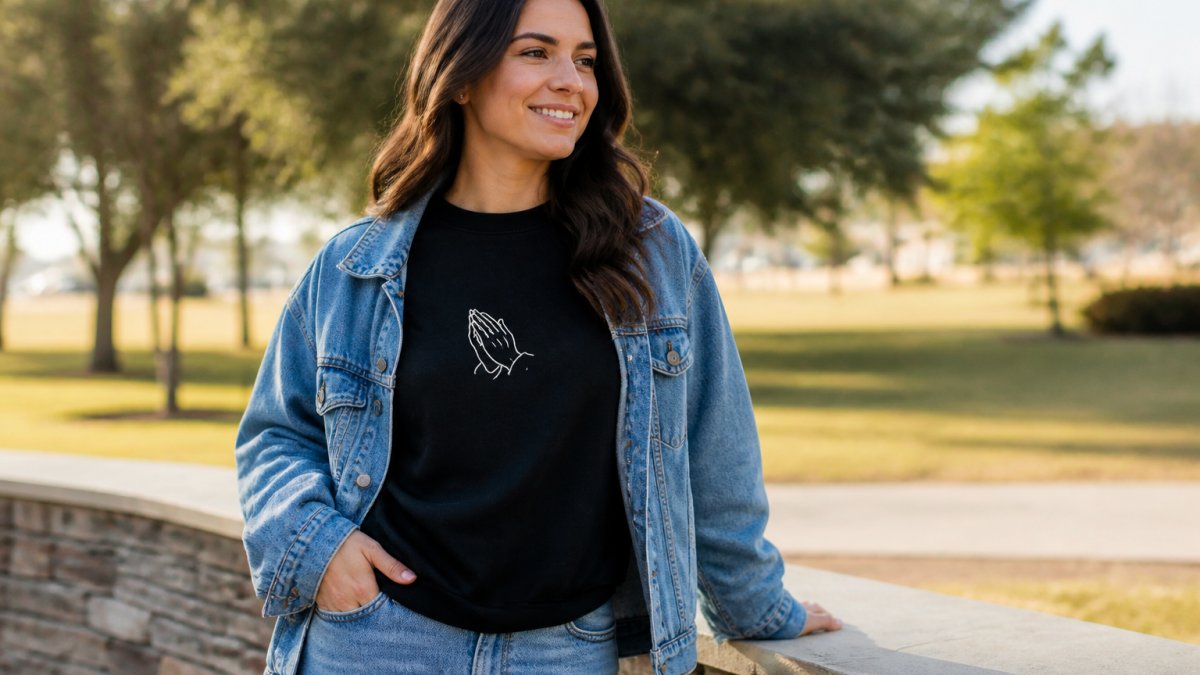 Woman wearing a black minimalist prayer sweatshirt layered with a denim jacket, styled casually with jeans for everyday wear in a bright, natural outdoor setting
