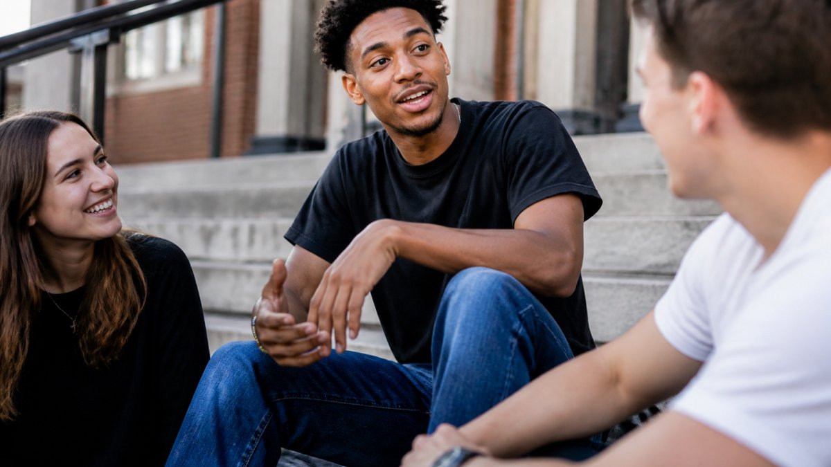 A young adult wearing a bold graphic tee sitting on outdoor steps with friends, mid-conversation, natural daylight, casual streetwear setting