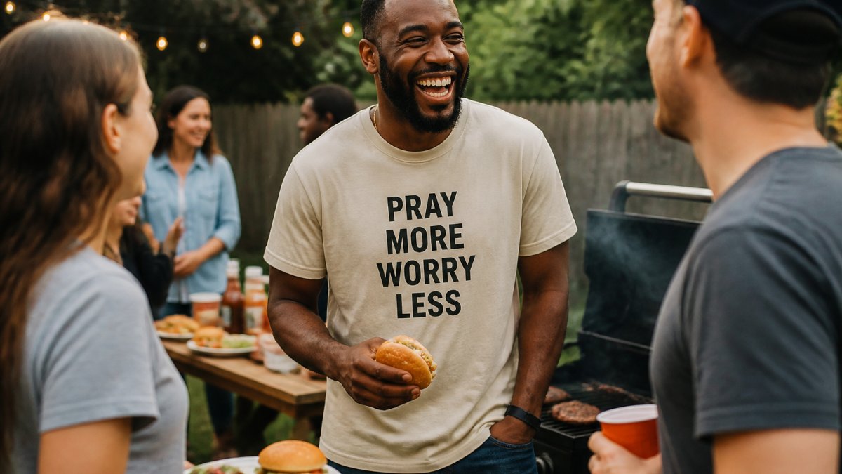A person in a relaxed-fit graphic tee laughing with friends at a backyard cookout, the shirt's printed phrase visible but the mood casual and unposed