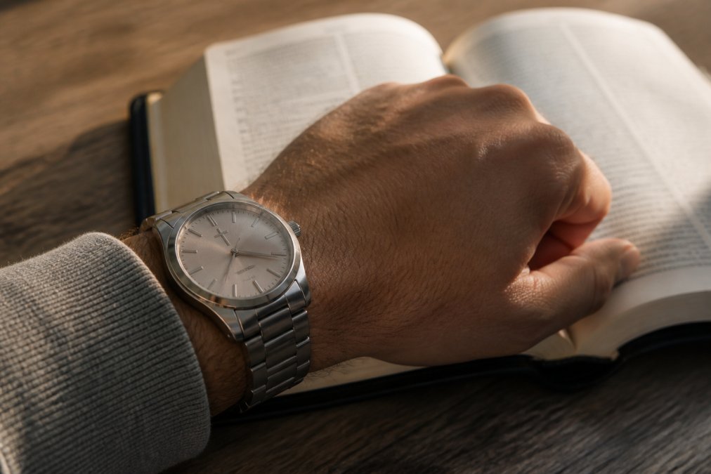Close-up of a man's wrist wearing a silver watch with a small cross on the dial, resting on an open Bible, soft natural light