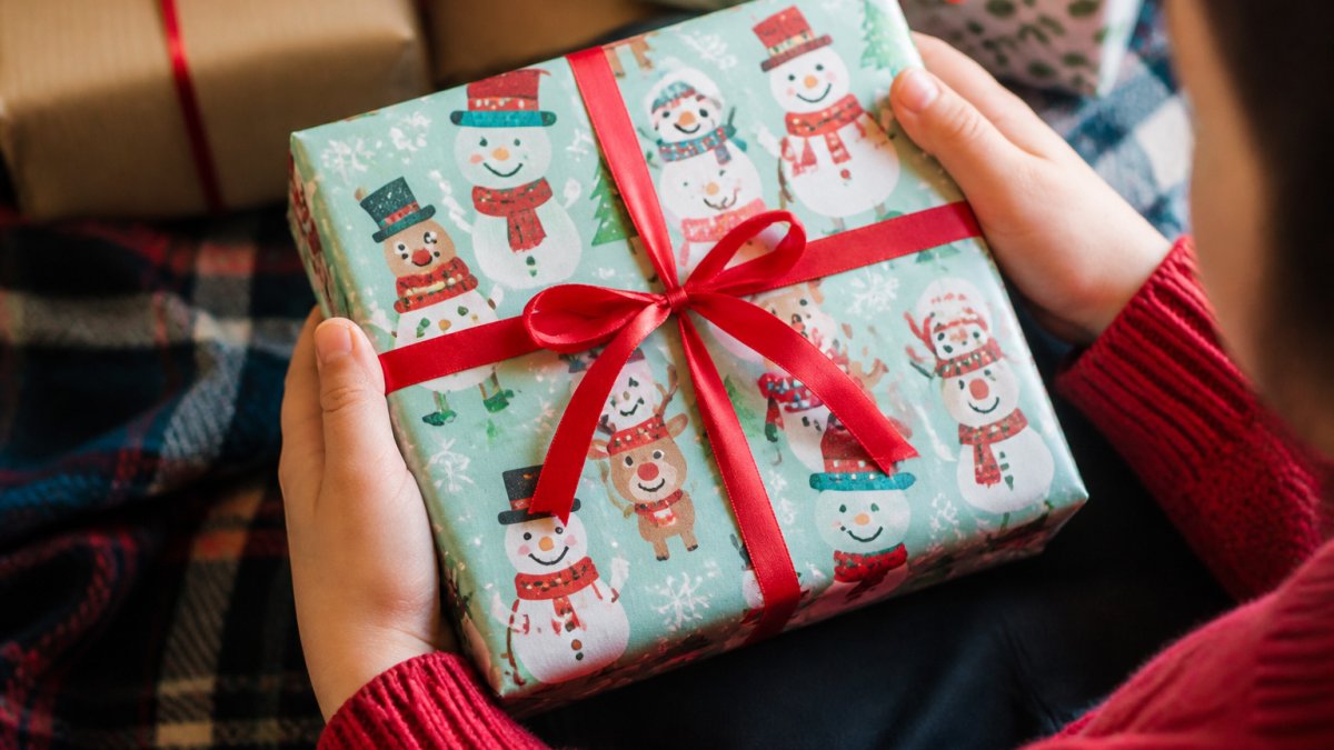 Child's hands holding a wrapped gift covered in cheerful cartoon snowman and reindeer designs, showing the festive pattern and paper texture in natural daylight