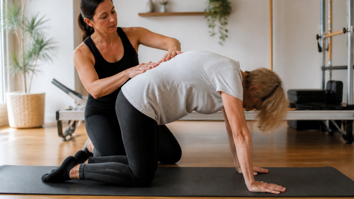 A Pilates instructor guiding a middle-aged adult through a gentle spinal articulation exercise on a mat in a calm, well-lit studio, focusing on controlled movement and posture