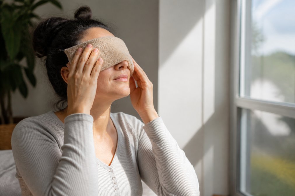 Person applying a warm compress to their closed eyes at home, sitting by a window with natural light streaming across their face