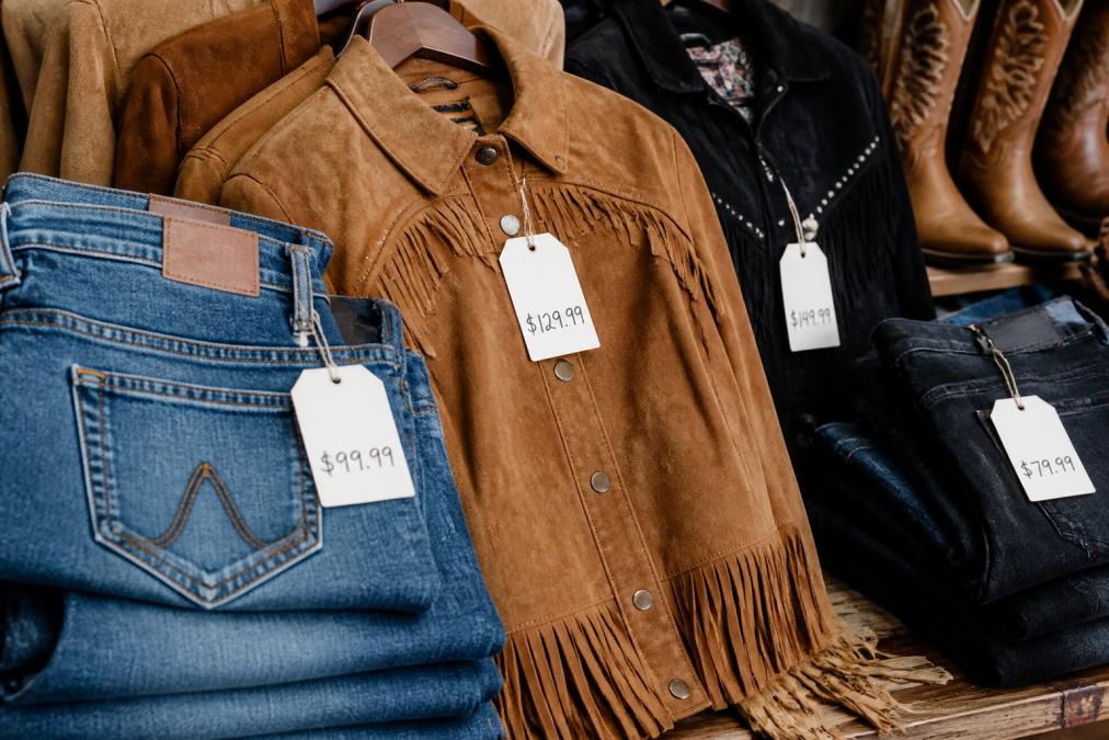 Close-up of western apparel items including quality denim jeans and fringe-trimmed jackets displayed with visible price tags, showing textile details and craftsmanship in natural lighting