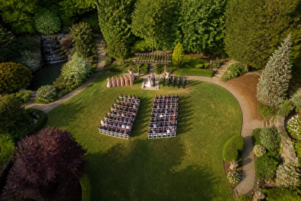 Aerial view of a wedding ceremony site with bride and groom visible from approximately 150 feet altitude, white chairs arranged in rows, guests seated, manicured garden landscaping surrounding the venue, natural afternoon lighting casting soft shadows across the grounds