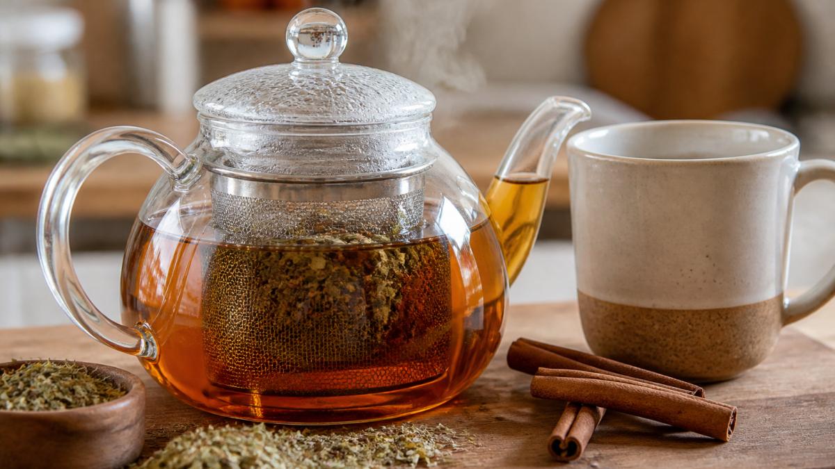 Close-up of a glass teapot filled with amber herbal tea, loose cinnamon sticks and dried oregano leaves steeping in an infuser basket, steam rising, on a wooden kitchen counter with a ceramic mug beside it
