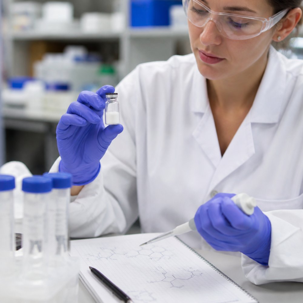 A close-up of a laboratory researcher's gloved hands holding a small vial of white lyophilized powder next to a pipette and a notebook with molecular sketches, in a bright, clean lab setting