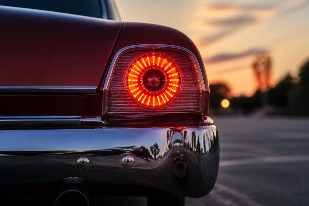 Close-up of a restored classic car tail light with bright red LED sequential lighting glowing at dusk, showing the contrast between the vintage chrome bumper and modern illumination