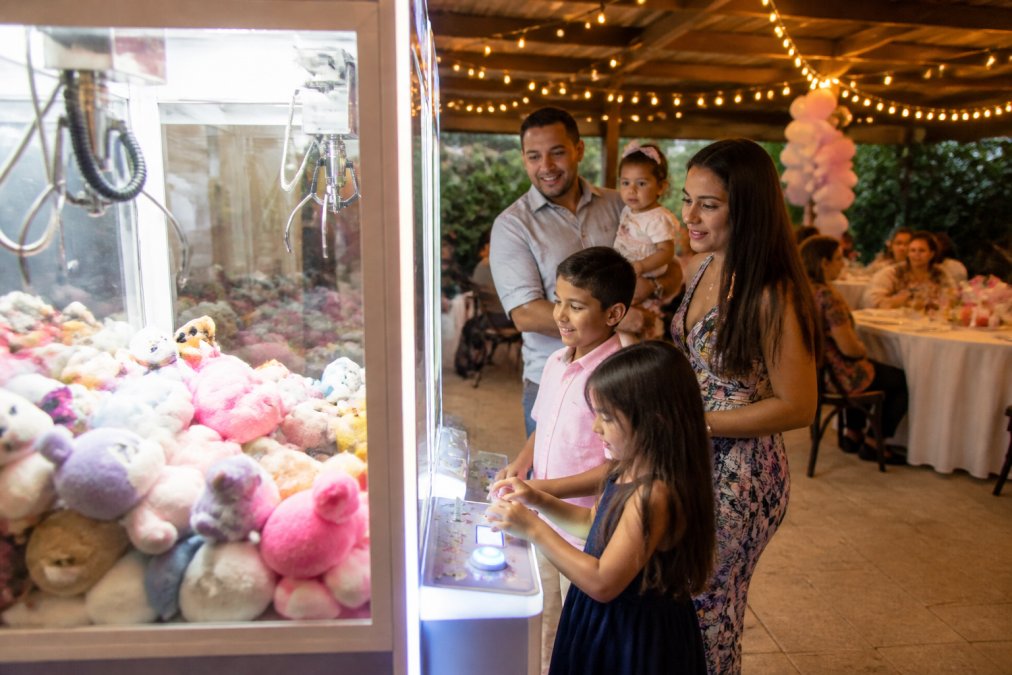 A family at a birthday party watching children play a lit claw machine filled with plush toys in a decorated event space, warm string lights overhead
