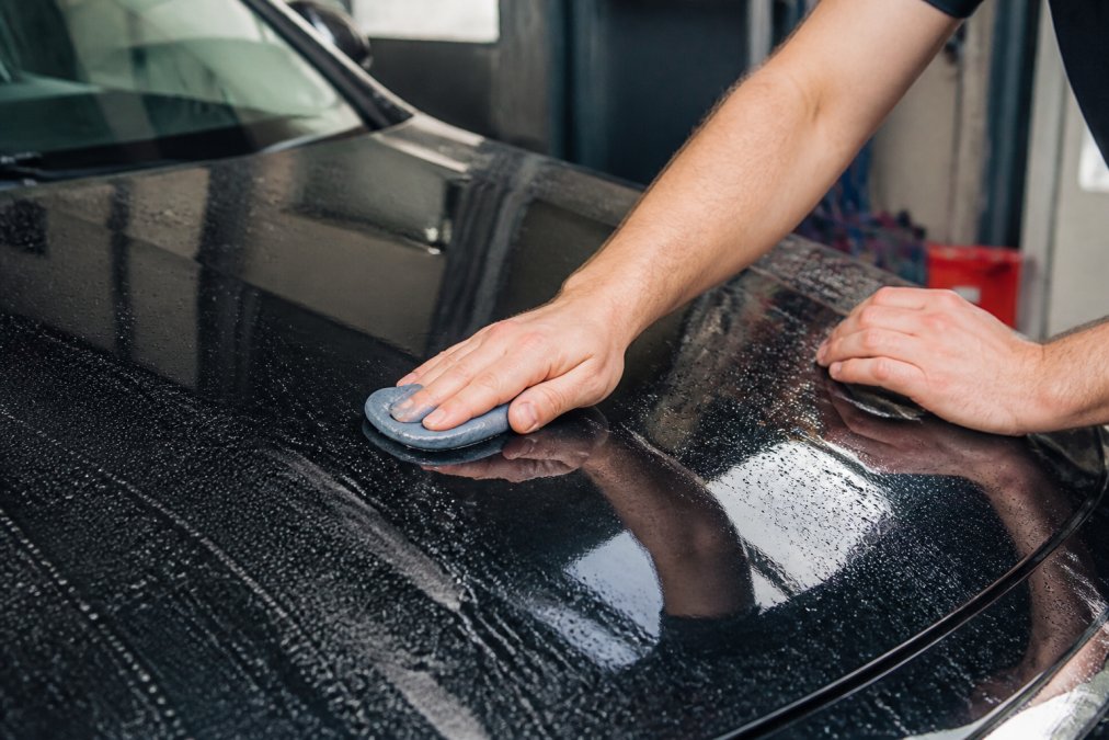 Handen die voorzichtig een medium clay bar over een natte, glimmende autolak bewegen met zichtbare spray-lubricant, onder helder daglicht in een detailwerkplaats
