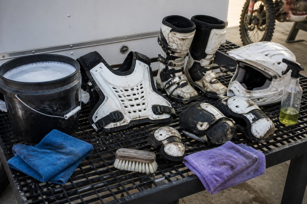 Motocross rider's protective gear laid out on a wash rack with bucket of soapy water, microfiber towels, soft brush, and degreaser bottle nearby, showing preparation for post-ride maintenance