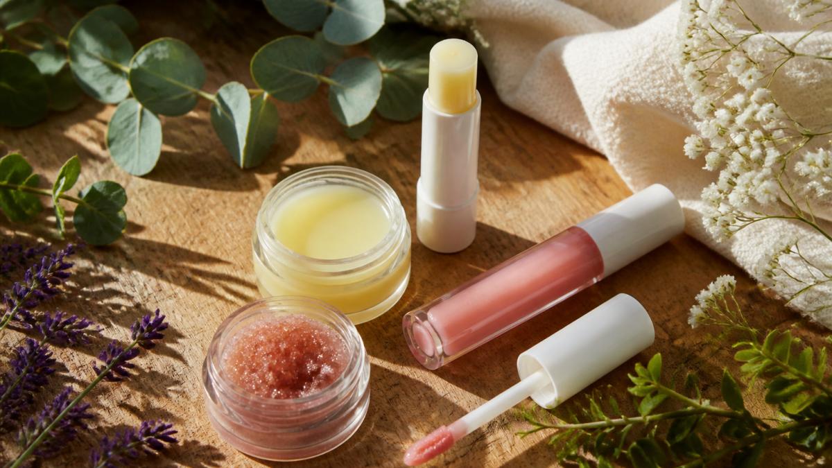 Lip balm, scrub, and gloss arranged with fresh botanicals on a natural wood surface in soft morning light