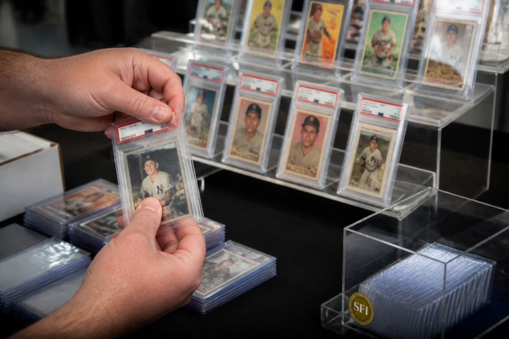 A dealer's hand placing a graded PSA slab inside a clear, snug-fit protective sleeve at a well-lit card show table, with other graded cards arranged neatly on tiered acrylic risers in the background