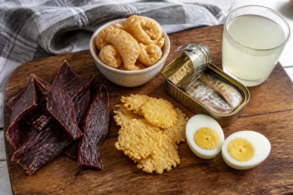 A flat-lay photo on a rustic wooden cutting board showing clean carnivore snacks: beef jerky strips, pork rinds in a small ceramic bowl, cheese crisps, an open tin of sardines, and two hard-boiled eggs, with a glass of electrolyte drink beside them, natural window lighting