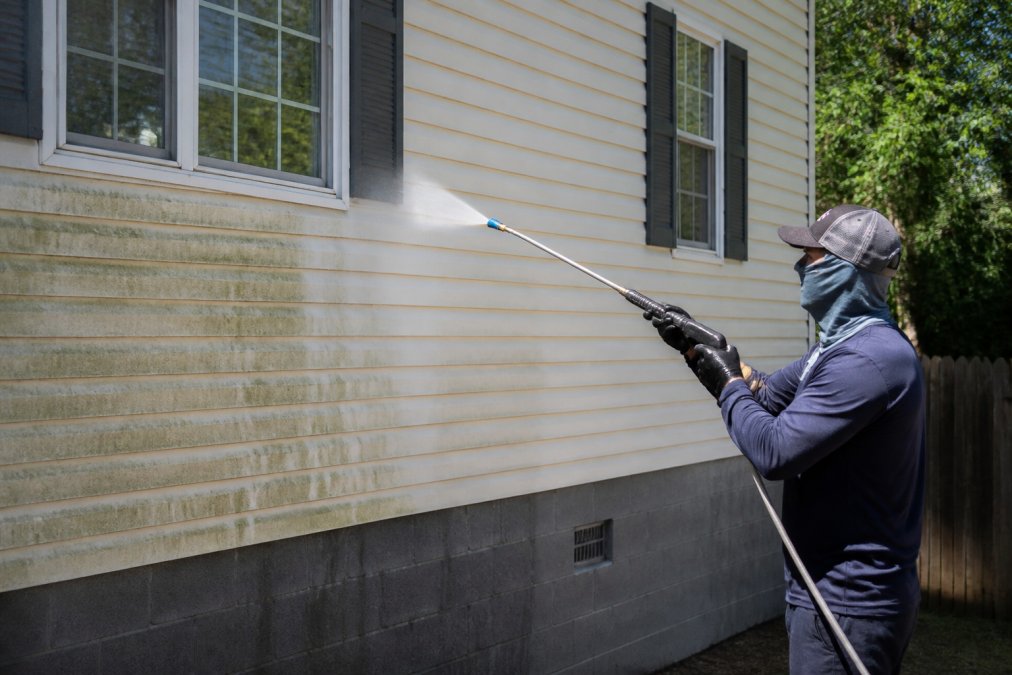 A technician in protective gear applying a low-pressure soft wash solution to residential vinyl siding with a long wand, showing a gentle spray angle under bright daylight
