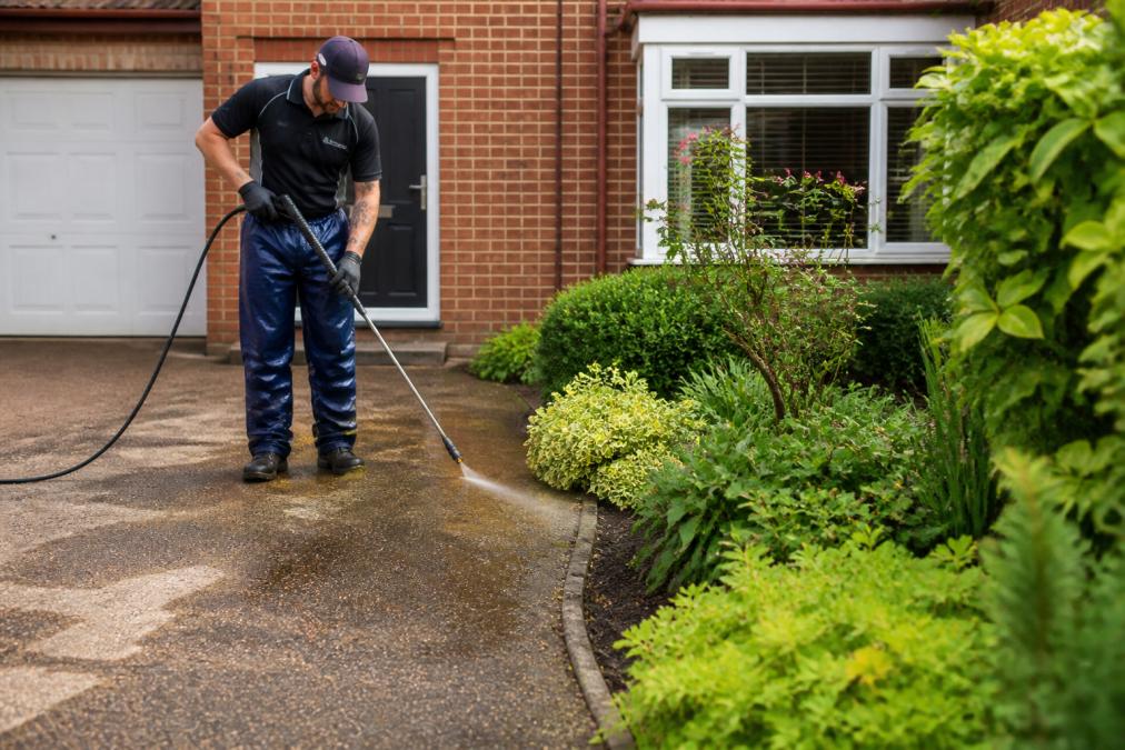 Professional demonstrating soft washing technique on residential driveway with careful attention to garden borders and plants, natural daylight