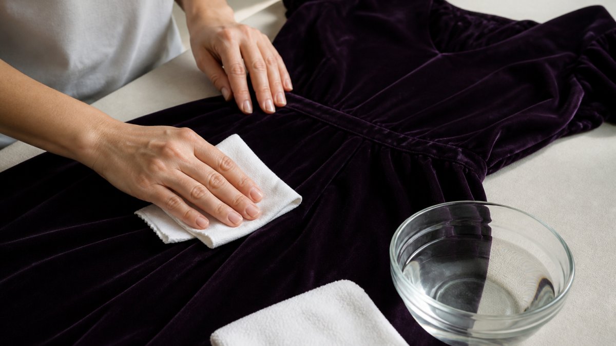 Close-up of hands gently blotting a dark velvet dress with a clean white microfiber cloth on a flat surface, with a small bowl of cool water nearby