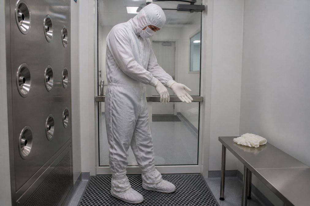 Cleanroom technician in full gowning procedure wearing sterile white suit, hood, gloves, and booties while performing step-by-step entry into controlled environment with proper hand positioning