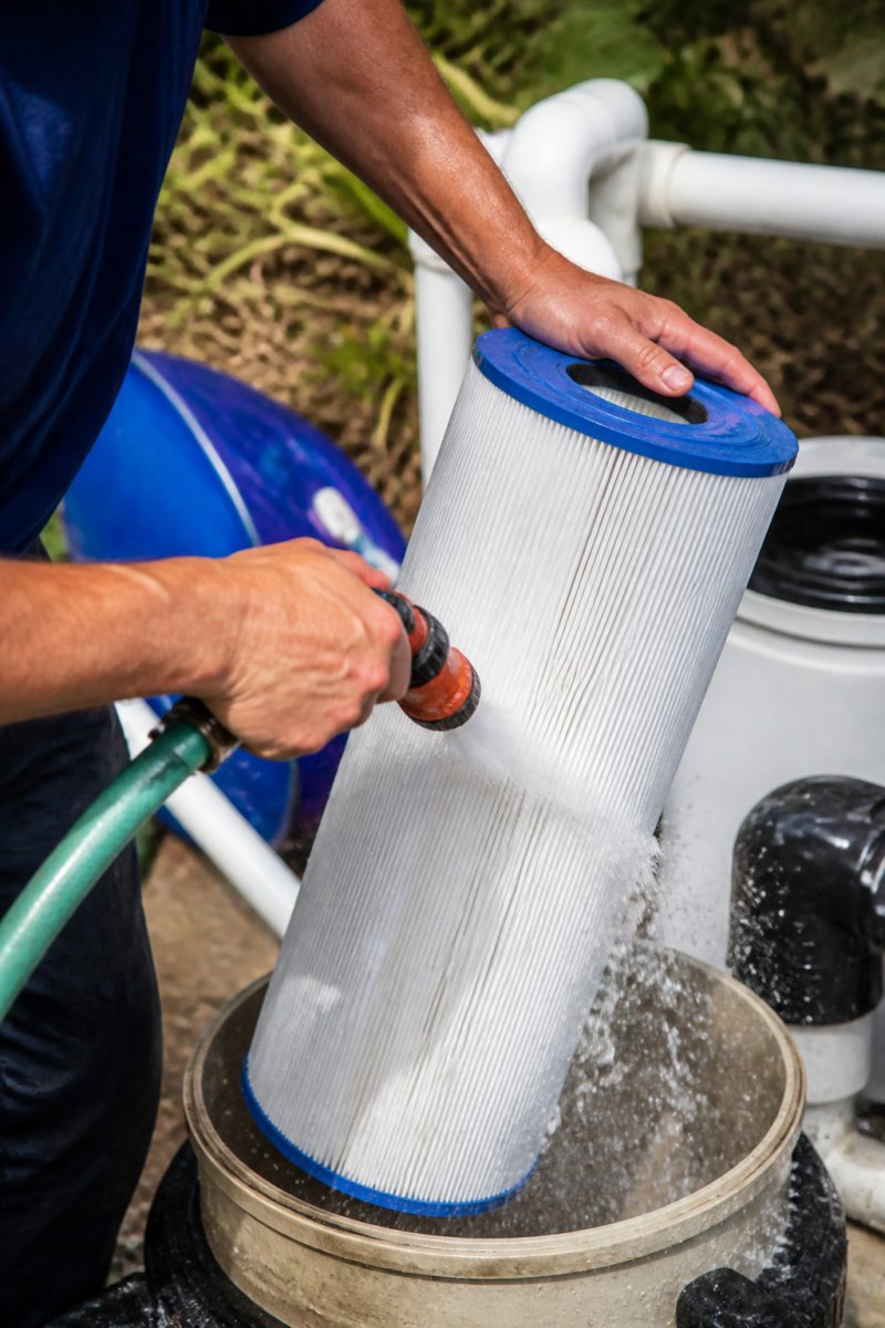 Close-up of a pool filter cartridge being rinsed with a garden hose, showing water spraying over the pleated filter element in bright daylight