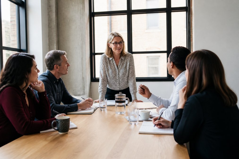 A trainer facilitating a small group discussion in a bright modern meeting room, participants engaged and taking notes, natural daylight through large windows