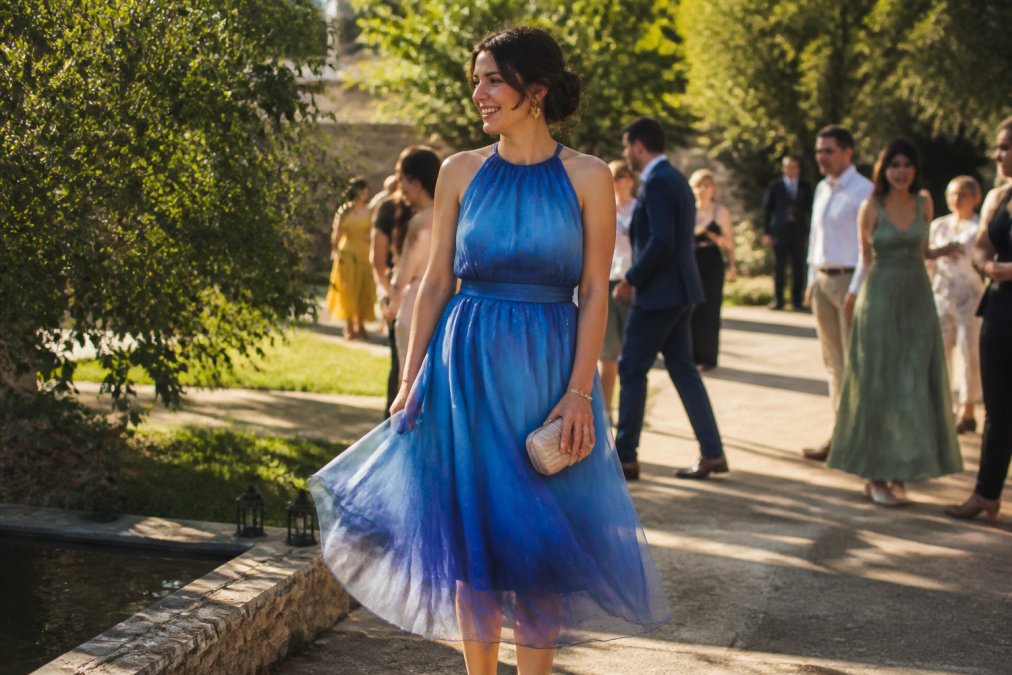 Woman wearing a jewel-toned blue ombré organza midi dress at an outdoor garden wedding, standing in natural sunlight with the metallic thread catching light, showcasing elegant drape and movement with minimal accessories