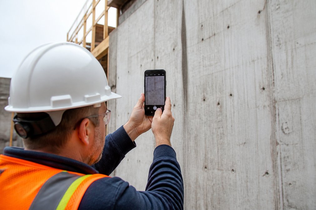 A field inspector in a hard hat and high-visibility vest holding an Android smartphone up to photograph a concrete wall at a construction site, with the app interface visible on screen showing a timestamped photo