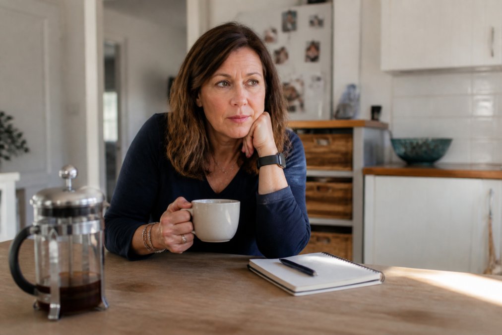 A woman in her mid-40s sitting at a kitchen table in the morning, holding a cup of tea, looking tired but determined, with a notebook and pen beside her