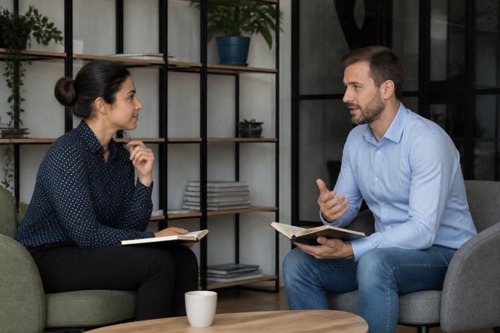 Professional coach and client seated across from each other in a modern office setting with natural light, both holding notebooks, engaged in active listening with focused attention