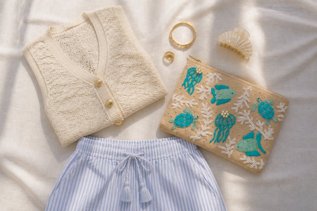Styled flat lay of coastal vacation outfit pieces arranged on light cream linen background including striped shorts, Coastal Charm Sweater Vest with visible gold shell buttons, Serenity Shell Ring and Bangle, seashell hair claw clip, and Natural Coastal Luxe Clutch, with soft natural window lighting