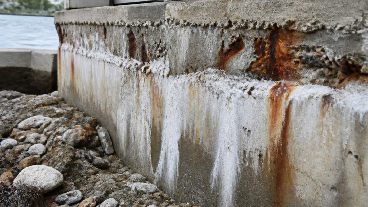 Close-up of concrete foundation showing visible spalling, rust staining, and white salt efflorescence deposits on a waterfront building exposed to salt spray