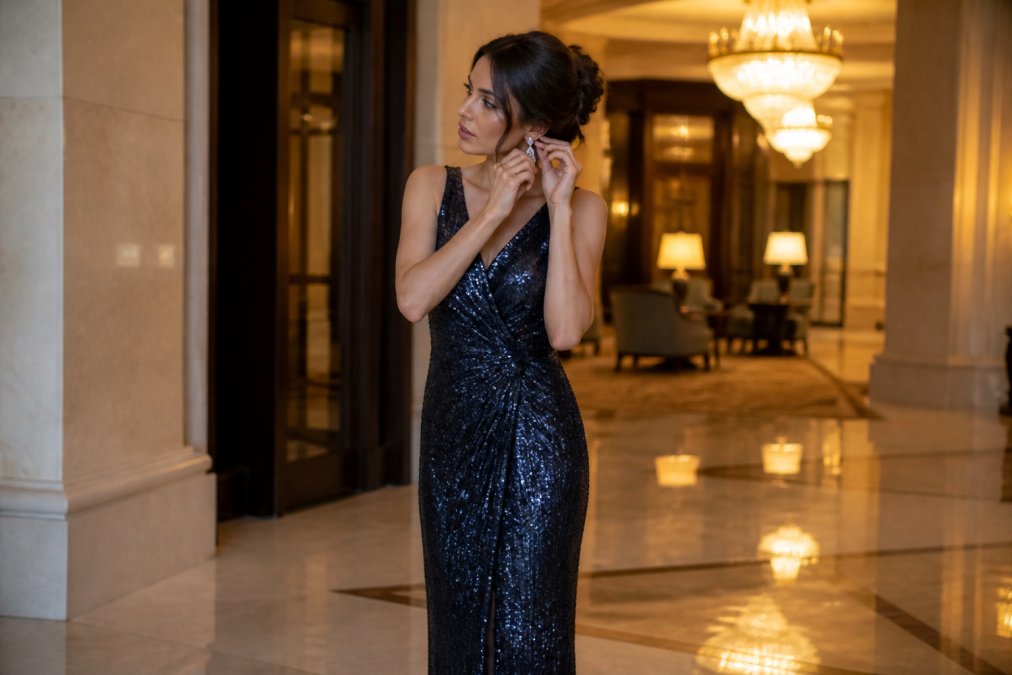 A woman in a floor-length sequinned evening gown standing in a softly lit hotel ballroom foyer, adjusting a drop earring, with marble floors and warm chandelier light behind her