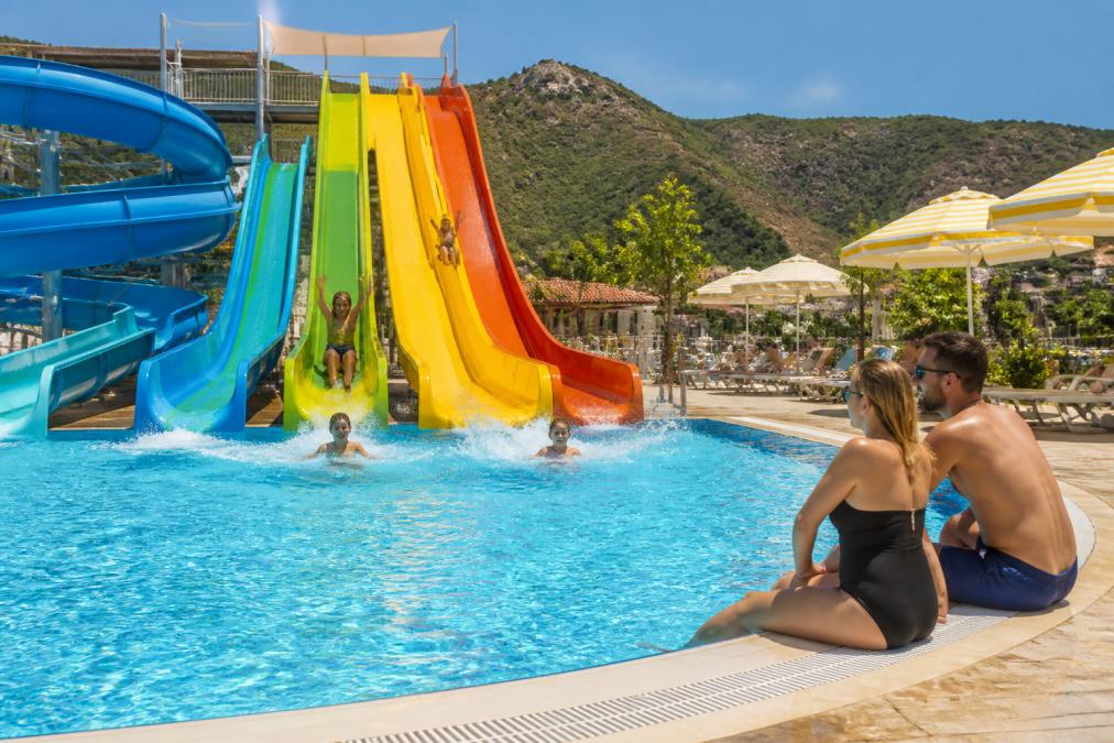 Orka World Waterpark'ta çocuklar renkli su kaydıraklarından kayıyor, ebeveynler yan tarafta izliyor, güneşli bir gün, havuzun etrafında şemsiyeler