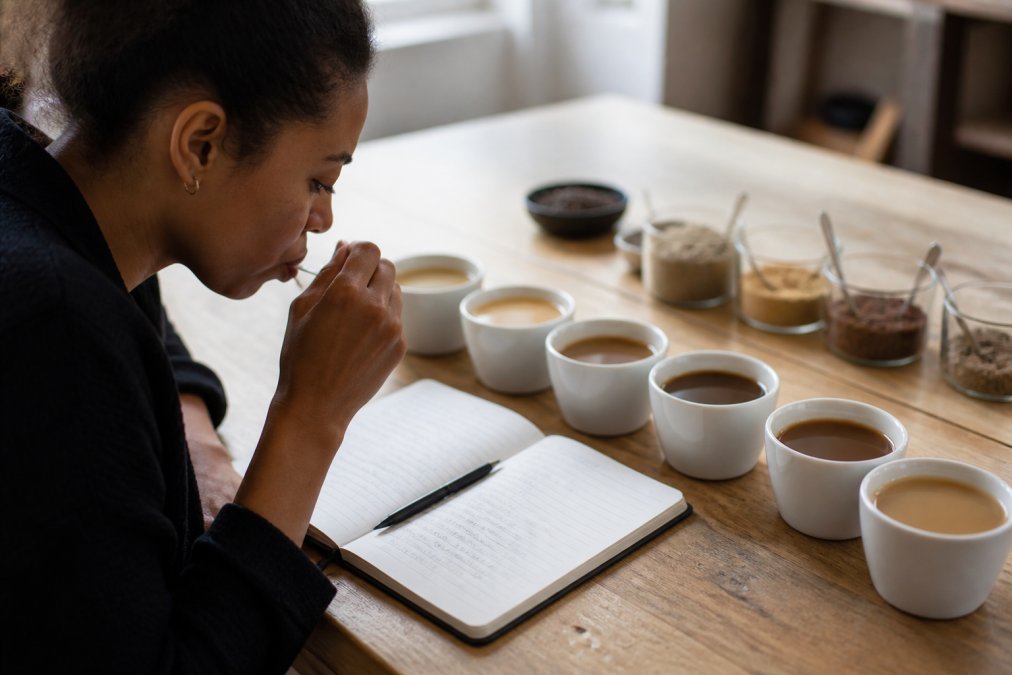 Close-up of someone tasting and evaluating different coffee alternative beverages in white cups, with tasting notes visible on paper nearby