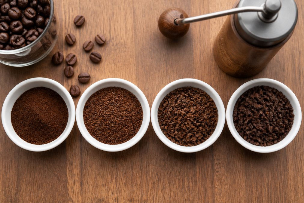 Overhead flat-lay of four small white dishes on a wooden counter, each holding a different coffee grind texture from fine espresso powder to coarse French press chunks, with a hand grinder and whole beans visible at the edge of the frame