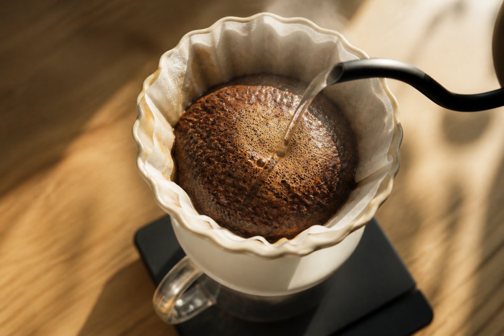 Overhead close-up of a ceramic pour over dripper sitting on a digital scale, hot water poured over dark coffee grounds that bubble and swell into a thick domed crust, steam rising in soft morning light