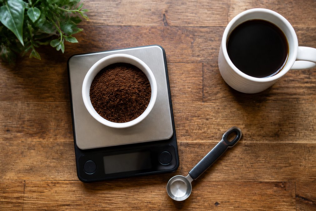 Overhead shot of a kitchen scale with a small bowl of coffee grounds, a tablespoon measure, and a mug of brewed coffee on a wooden countertop