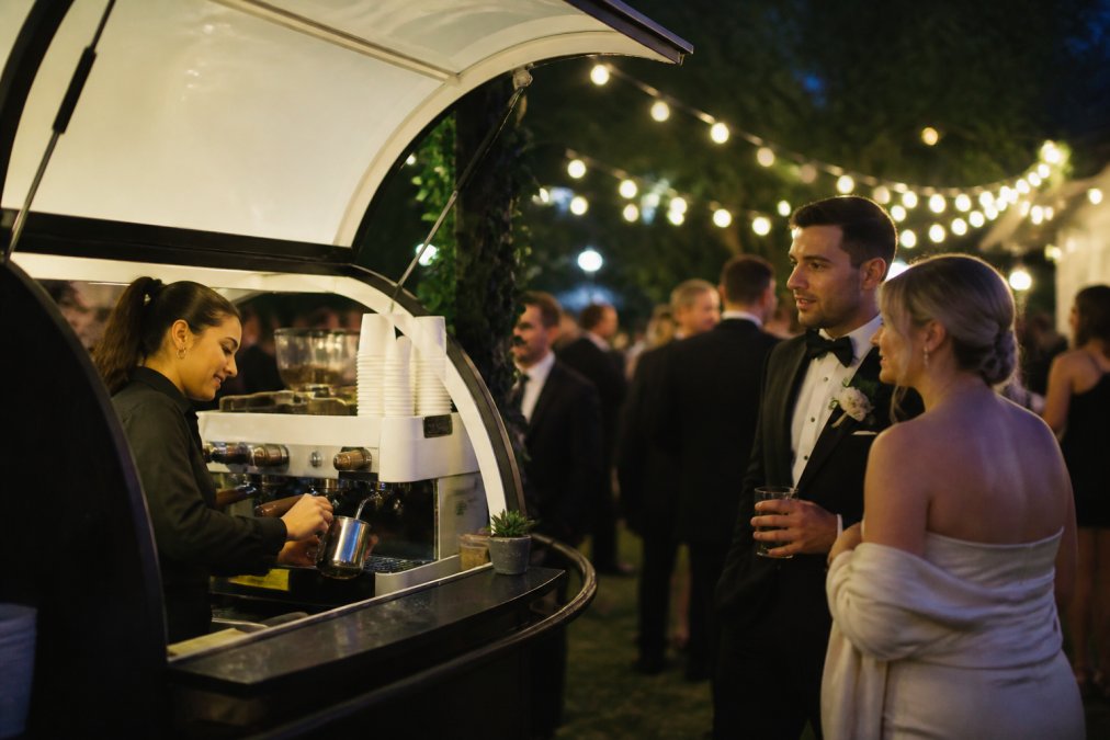Professional barista preparing specialty espresso drinks at a mobile coffee cart during an outdoor wedding reception, with guests in formal attire and garden lighting in the background