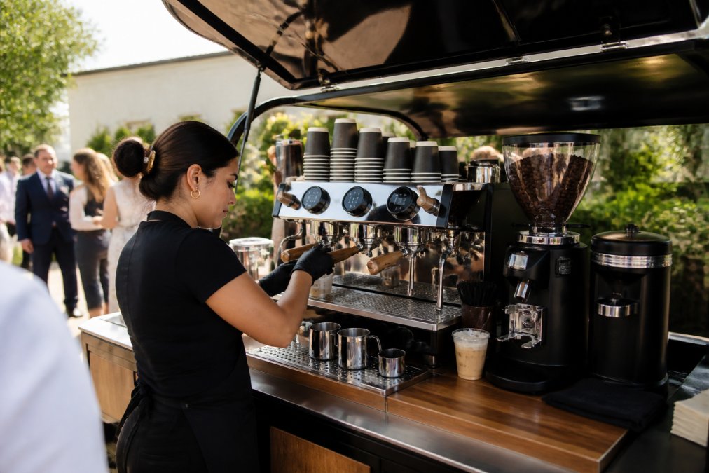 Professional barista preparing specialty espresso drinks at a fully equipped mobile coffee cart with dual boiler espresso machine, commercial grinder, and milk steamer visible, mid-service with event guests waiting in background, natural outdoor lighting