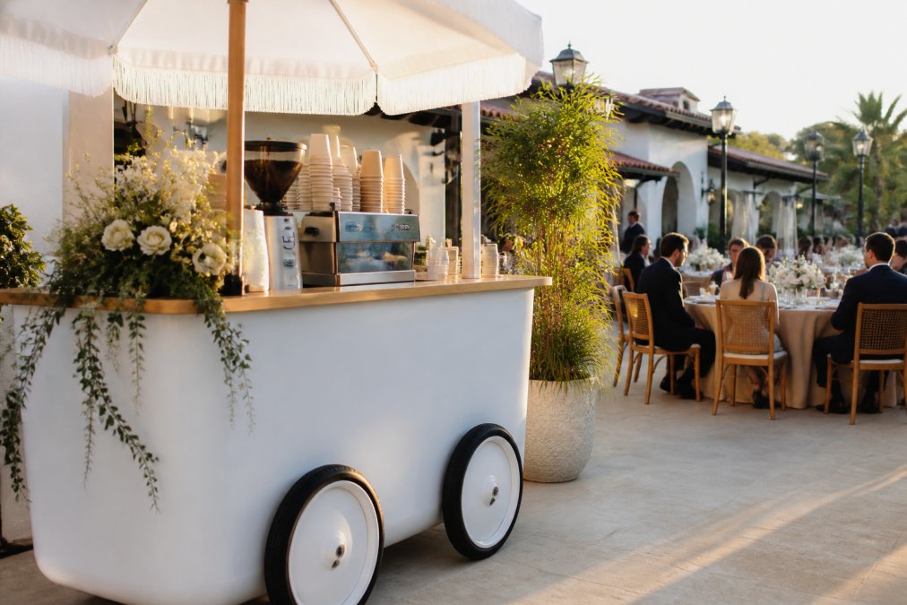 Custom-branded mobile espresso cart at an upscale outdoor wedding venue with elegant signage and professional setup, positioned near event seating with guests enjoying drinks in background, soft natural lighting
