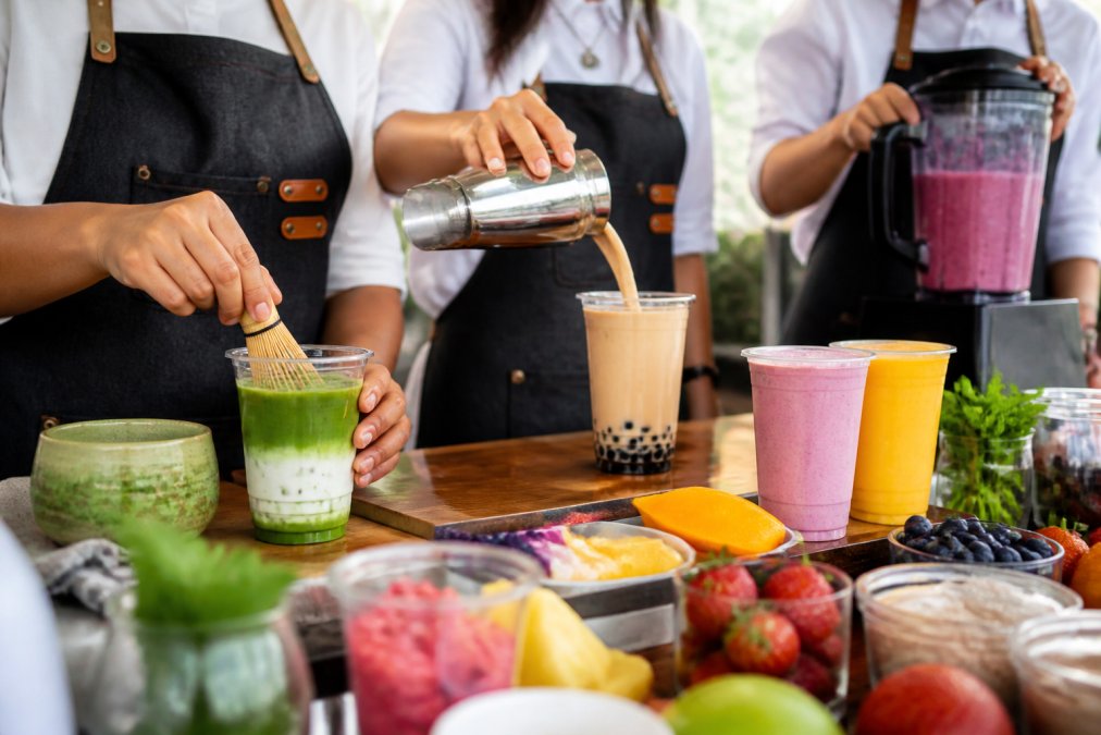 Close-up of hands preparing specialty drinks at a vibrant beverage station, matcha being whisked into a creamy latte, boba tea being poured with tapioca pearls visible, fresh smoothies being blended with colorful fruits and berries, with an array of colorful cups and fresh garnishes arranged in the foreground