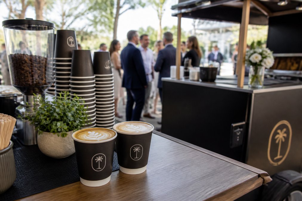 Close-up of custom branded coffee cups featuring corporate logo and specialty latte art at an upscale event, with branded signage visible on the mobile coffee cart in soft natural lighting