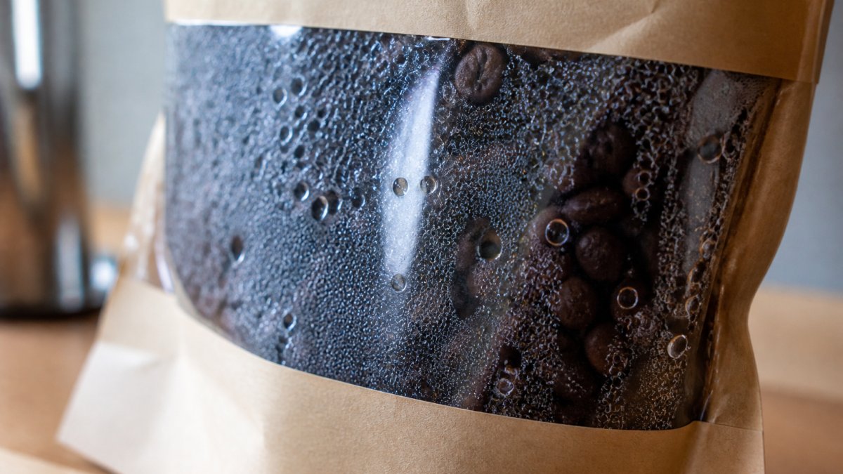 Close-up of freshly roasted dark coffee beans in a sealed kraft bag with visible moisture beads and condensation on the interior surface, showing active degassing in progress