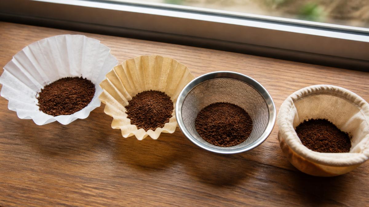 Close-up arrangement of different coffee filters displayed side-by-side on a wooden surface: white bleached paper filter, tan unbleached paper filter, stainless steel mesh filter, and organic cotton cloth filter, with fresh medium-roast coffee grounds visible on or near each, natural window lighting